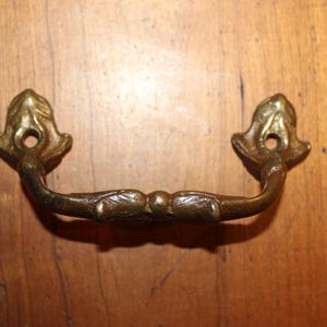 May include: A gold-toned metal drawer pull with a decorative leaf design. The pull has a simple, elegant style.