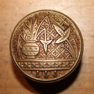 May include: A detailed, antique brass button with an embossed design. The central motif features a vase of flowers, a stylized bird, and geometric patterns. The button is circular and has a detailed border, showcasing a vintage aesthetic.