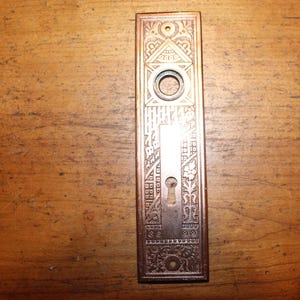 May include: An antique bronze door plate with intricate geometric and floral patterns. The rectangular plate features a keyhole, a circular opening, and detailed engravings, showcasing a vintage aesthetic. The plate is set against a wooden surface.