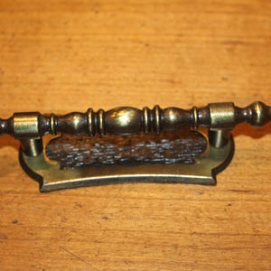 May include: A decorative antique brass drawer pull with a dark brown center. The pull has a detailed, ornate design with a curved base and a cylindrical handle. The hardware is likely for a cabinet or dresser.