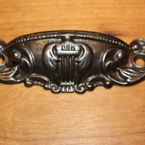 May include: A black metal drawer pull with an ornate design featuring a lyre in the center. The pull has two holes for mounting.