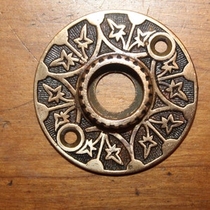 May include: A round, brass door knob escutcheon with a decorative design of stars and leaves. The escutcheon has a hole in the center for the knob.