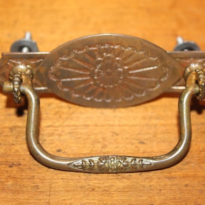 May include: Antique brass drawer pull with a decorative oval center featuring a sunburst design. The handle is a curved, U-shaped design with decorative detailing. Includes mounting hardware.
