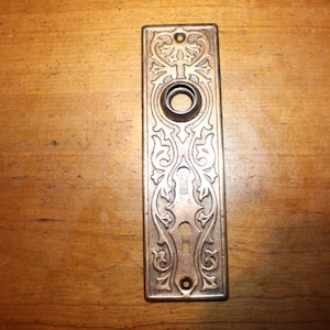 May include: An antique bronze-colored door plate with ornate floral and scrollwork designs. The rectangular plate features a central keyhole and a circular opening for a doorknob. Two screw holes are visible at the top and bottom.