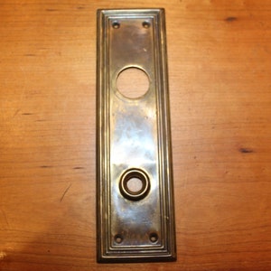 May include: A brass door plate with a rectangular shape and a decorative border. It has two round holes, one larger than the other, for a door knob and a keyhole.