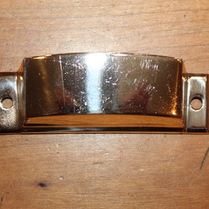 May include: A gold-toned metal drawer pull with a curved top and two holes for mounting.