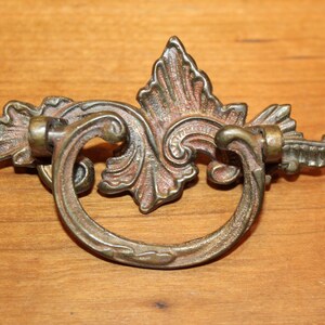 May include: Ornate brass drawer pull with a decorative, scrolled design. The pull has a ring shape and is attached to a decorative, leaf-shaped backplate.