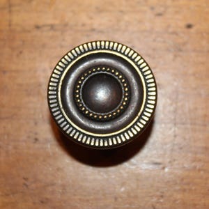 May include: A round, brass cabinet knob with a concentric circle design. The knob has a dark brown finish with a gold trim.