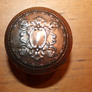 May include: A round, silver-colored door knob with an ornate design. The knob has a raised, circular center with a floral pattern.