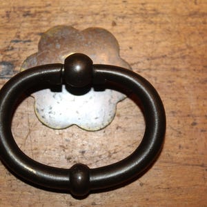 May include: A black metal ring pull handle with a decorative silver flower-shaped backplate. The handle is attached to a wooden surface.