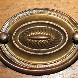 May include: Antique brass drawer pull with an oval, ornate design. The center features a detailed, swirling pattern, and the pull has two rounded knobs on either side. The brass has a warm, aged patina.