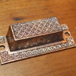 May include: A rectangular, antique-style, brass door pull with a decorative, geometric pattern. The pull has two holes for mounting.