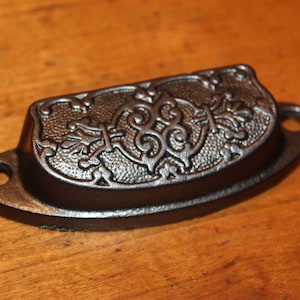 May include: A dark bronze-colored drawer pull with an ornate, raised floral and scroll design. The handle has a curved shape with two mounting holes on either end. The hardware is likely made of cast iron or a similar metal.