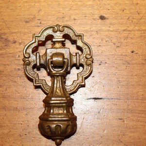May include: Ornate antique brass drawer pull with a decorative, openwork design. The pull features a detailed, scrolling pattern and a central handle. The hardware is set against a wooden surface.