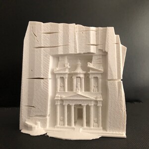 May include: A white 3D-printed sculpture of the Treasury at Petra, Jordan. The sculpture is carved into a block with a rough, textured exterior, and the facade of the Treasury is intricately detailed with columns and architectural elements.