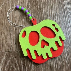 May include: A green and red wooden apple ornament with a skull design. The apple has a green dripping effect and is attached to a white string with purple and green beads.