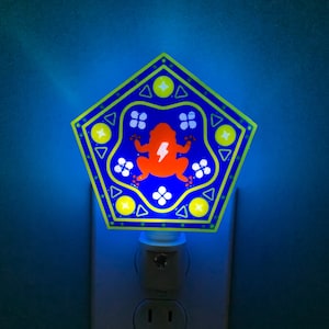 May include: A blue and yellow nightlight shaped like a pentagon with a frog inside. The frog is orange and has a lightning bolt on its chest. The nightlight has a white plug.