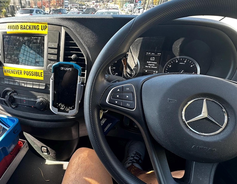 Puede incluir: Interior de un veh&iacute;culo con volante negro y logotipo de Mercedes-Benz. Un dispositivo GPS est&aacute; montado cerca de la pantalla de infoentretenimiento. Pegatinas amarillas con los textos "AVOID BACKING UP" y "WHENEVER POSSIBLE".