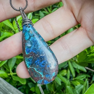 May include: A teardrop-shaped pendant featuring a polished stone with deep blue, turquoise, and dark brown marbled patterns. The stone is secured by a dark, oxidized silver wire wrap attached to a matching twisted metal ring and chain necklace.