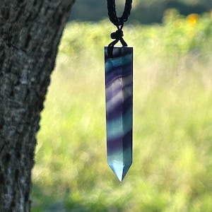 May include: A purple and blue fluorite crystal pendant on a black cord necklace. The crystal is faceted and has a pointed tip. The pendant is hanging against a blurred background of green foliage.