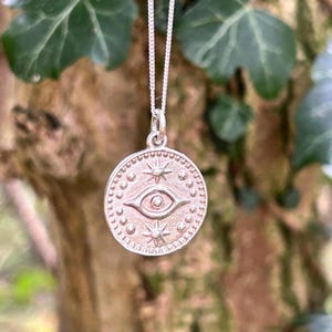 May include: Silver necklace with a round pendant featuring a stylised eye with stars and dots around it.