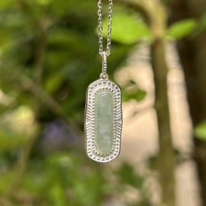 Op de afbeelding: Een zilveren ketting met een rechthoekige hanger met een lichtgroene edelsteen omringd door een decoratieve zilveren rand.