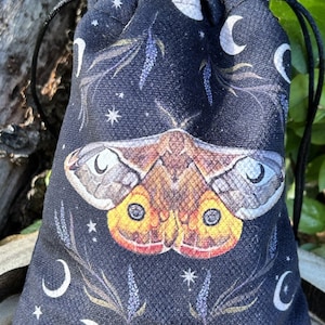 Pode incluir: Uma bolsa de cordão preto com um padrão de lua e estrelas. A bolsa apresenta uma grande mariposa colorida com asas amarelas e laranja. A bolsa é perfeita para guardar pequenos objetos como cristais, joias ou cartas de tarô.