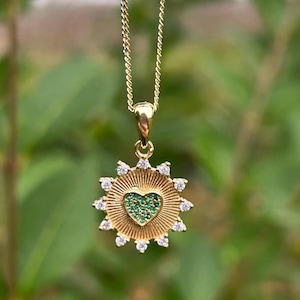 Puede incluir: Un collar de oro con un colgante que presenta un corazón verde rodeado de un diseño de sol con piedras transparentes.