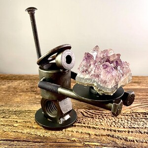 May include: A unique sculpture made from metal hardware, holding a cluster of amethyst crystals. The sculpture features bolts, nuts, and washers, with a textured, industrial aesthetic. The amethyst is a light purple color.