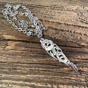 May include: A silver-colored pendant necklace with an ornate, elongated design. The pendant features intricate floral and scrollwork patterns. The necklace chain is also silver and has a delicate link design. The necklace is displayed on a rustic wooden surface.