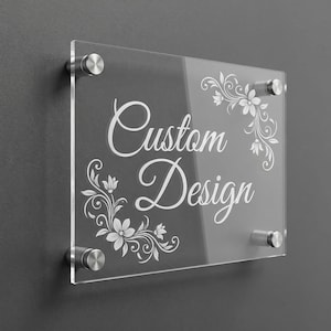 Könnte beinhalten: Transparente Acrylglas-Tafel mit dem Schriftzug "Custom Design" in elegantem Weiß. Florale Verzierungen schmücken die Ecken. An einer grauen Wand mit silbernen Abstandshaltern befestigt, für eine moderne und personalisierte Optik.