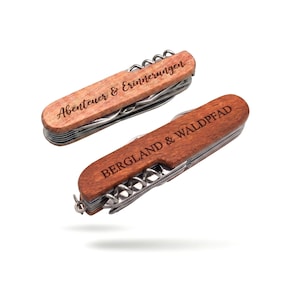 Puede incluir: Dos navajas suizas de madera con herramientas de acero inoxidable. Una navaja tiene el texto "Abenteuer & Erinnerungen" y la otra "BERGLAND & WALDPFAD" grabados en el mango de madera. Las navajas están sobre un fondo blanco.
