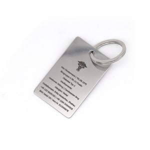 Könnte beinhalten: Ein silberner Schlüsselanhänger aus Metall mit einem medizinischen Symbol und gravierten Informationen. Der Schlüsselanhänger enthält den Text "Max Mustermann | 01.09.1975", "Blutgruppe A Rh (+) Positiv", "Diabetes Typ 2", "Medikamente:", "Metformin 500mg | Candesartan 8mg", "Allergien: Nickel", "Notfallkontakt: Erika Musterit, Ehefrau", "+49 123 5625132 | +49 1234 54344318", "BKK Mobil Oll | Vers. Nr. E12345678".