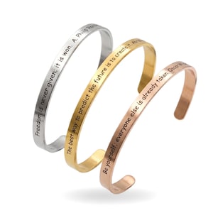 Op de afbeelding: Drie metalen manchetarmbanden in zilver, goud en roségoud. Elke armband heeft een inspirerende quote aan de buitenkant gegraveerd. De zilveren armband leest "Vrijheid wordt nooit gegeven; het wordt gewonnen". De gouden armband zegt "De beste manier om de toekomst te voorspellen is om deze te creëren". De roségouden armband zegt "Wees jezelf; iedereen is al bezet."
