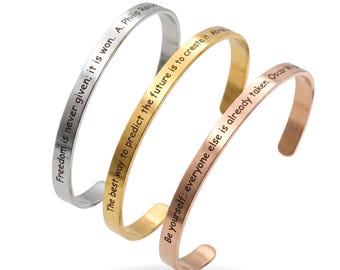 Op de afbeelding: Drie metalen manchetarmbanden in zilver, goud en roségoud. Elke armband heeft een inspirerende quote aan de buitenkant gegraveerd. De zilveren armband leest "Vrijheid wordt nooit gegeven; het wordt gewonnen". De gouden armband zegt "De beste manier om de toekomst te voorspellen is om deze te creëren". De roségouden armband zegt "Wees jezelf; iedereen is al bezet."
