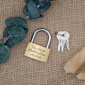 Può includere: Un lucchetto dorato con la scritta "Two Hearts Locked in Love - Australia" su uno sfondo di juta. Il lucchetto è accompagnato da tre chiavi.