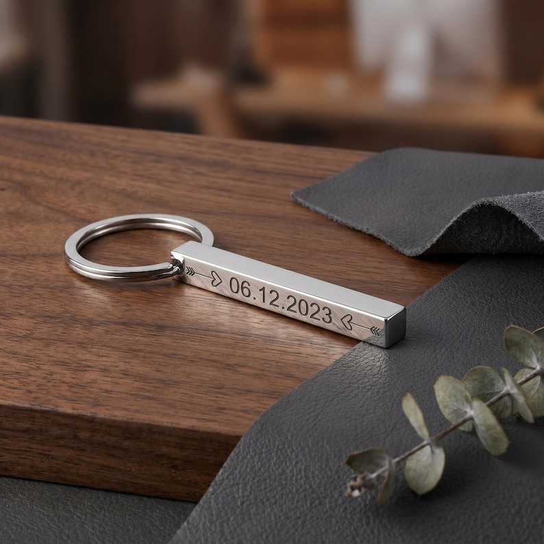 May include: A silver rectangular keychain with the date "06.12.2023" engraved on it, along with arrow and leaf designs. The keychain is on a wooden surface with a dark gray leather-like material and a sprig of greenery.