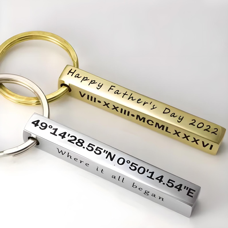 May include: Two keychains with rectangular pendants. One is gold-colored and reads "Happy Father's Day 2022" and Roman numerals. The other is silver and has coordinates and the phrase "Where it all began."