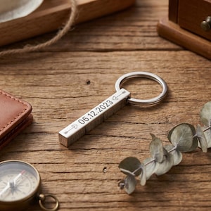 May include: A silver rectangular keychain with a ring, engraved with the date "06.12.2023" and arrows. The keychain is on a wooden surface, with a compass and a leather wallet in the background.
