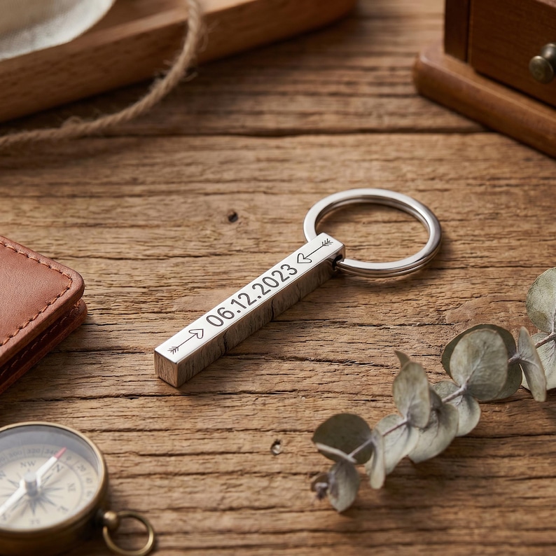 May include: A silver rectangular keychain with a ring, engraved with the date "06.12.2023" and arrows. The keychain is on a wooden surface, with a compass and a leather wallet in the background.