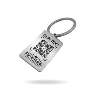 May include: A rectangular silver-toned metal keychain featuring a brushed finish and a circular split ring. The surface displays a laser-engraved QR code, the text DEIN TEXT, and the brand name GRAVURENKING with a small crown icon and the year 2018.