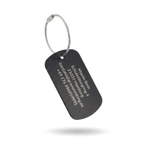 Peut inclure: Une étiquette de bagage noire avec une boucle en fil d'argent. L'étiquette a des coins arrondis et comporte du texte blanc, notamment un nom, une adresse et des coordonnées. L'étiquette est suspendue sur un fond blanc.