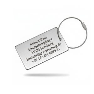 May include: A rectangular silver luggage tag with a wire loop. The tag has the name "Maxim Stein" and contact information printed in black. The tag is designed for easy identification of luggage.