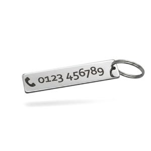 Pode incluir: Uma etiqueta retangular de chaveiro de metal prateado com um ícone de telefone e o número de telefone 0123 456789 gravado. A etiqueta é presa a um chaveiro de metal prateado. O fundo é branco.