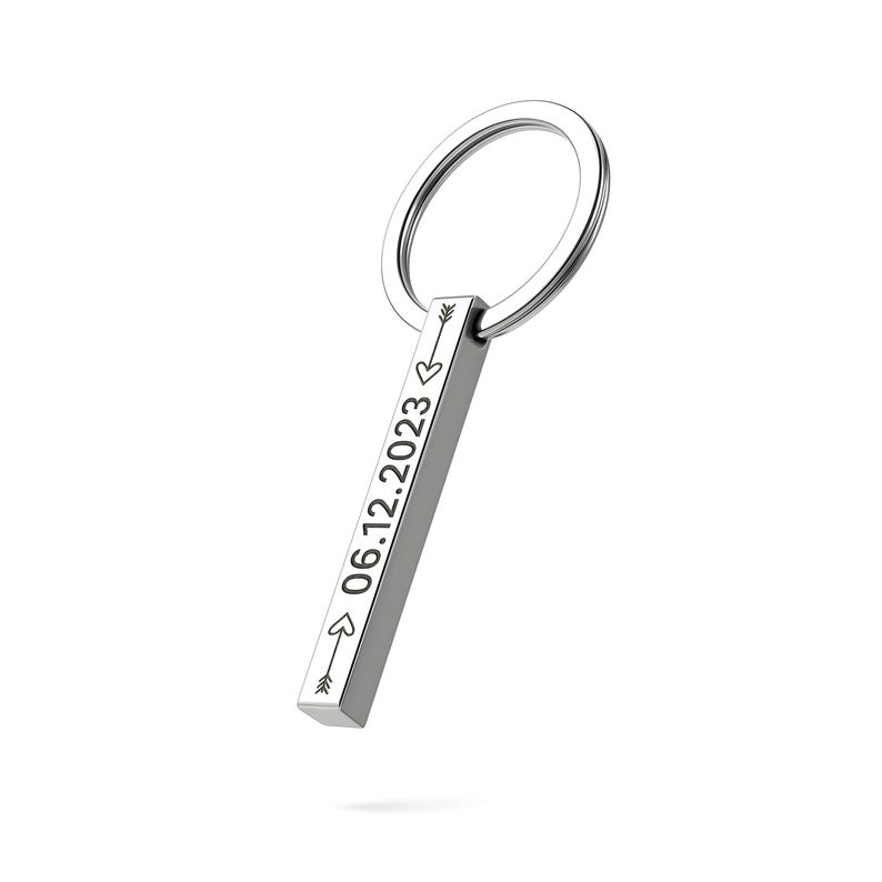 May include: A silver-colored keychain with a rectangular bar pendant. The bar is engraved with the date "06.12.2023" and arrow symbols. The keychain has a circular ring for attaching keys.