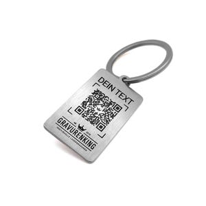 Pode incluir: Um porta-chaves retangular prateado com um código QR e o texto "DEIN TEXT" na parte superior. A parte inferior do porta-chaves tem o texto "GRAVURENKING" e um logótipo de coroa. O porta-chaves está preso a um anel prateado.
