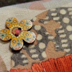 Könnte beinhalten: Ein cremefarbenes Stoffstück mit einem mehrfarbigen Blumenmuster, einem dunkelgrauen, strukturierten kreisförmigen Design und einem leuchtend orangefarbenen Fransenbesatz. Ein blumenförmiger Holzknopf mit gelbem, blauem und orangefarbenem Druck ist mit rosa Faden befestigt.