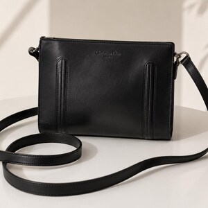 May include: A black rectangular crossbody bag featuring a smooth finish, a thin adjustable shoulder strap, and two vertical stitched panels on the front. The text Christian Dior Paris is embossed in the center of the upper portion of the bag.