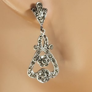 Puede incluir: Pendientes colgantes plateados con un diseño decorativo. Los pendientes presentan una forma floral con pequeños detalles oscuros. Los pendientes se muestran en una cabeza de maniquí.