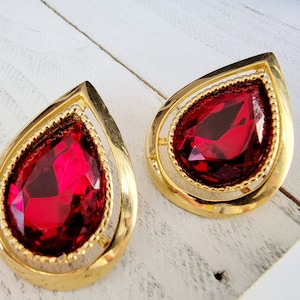 May include: Pair of gold-tone teardrop earrings with large, faceted, ruby-red center stones. The earrings have a detailed gold frame with a beaded inner edge. The earrings are displayed on a white wooden surface.